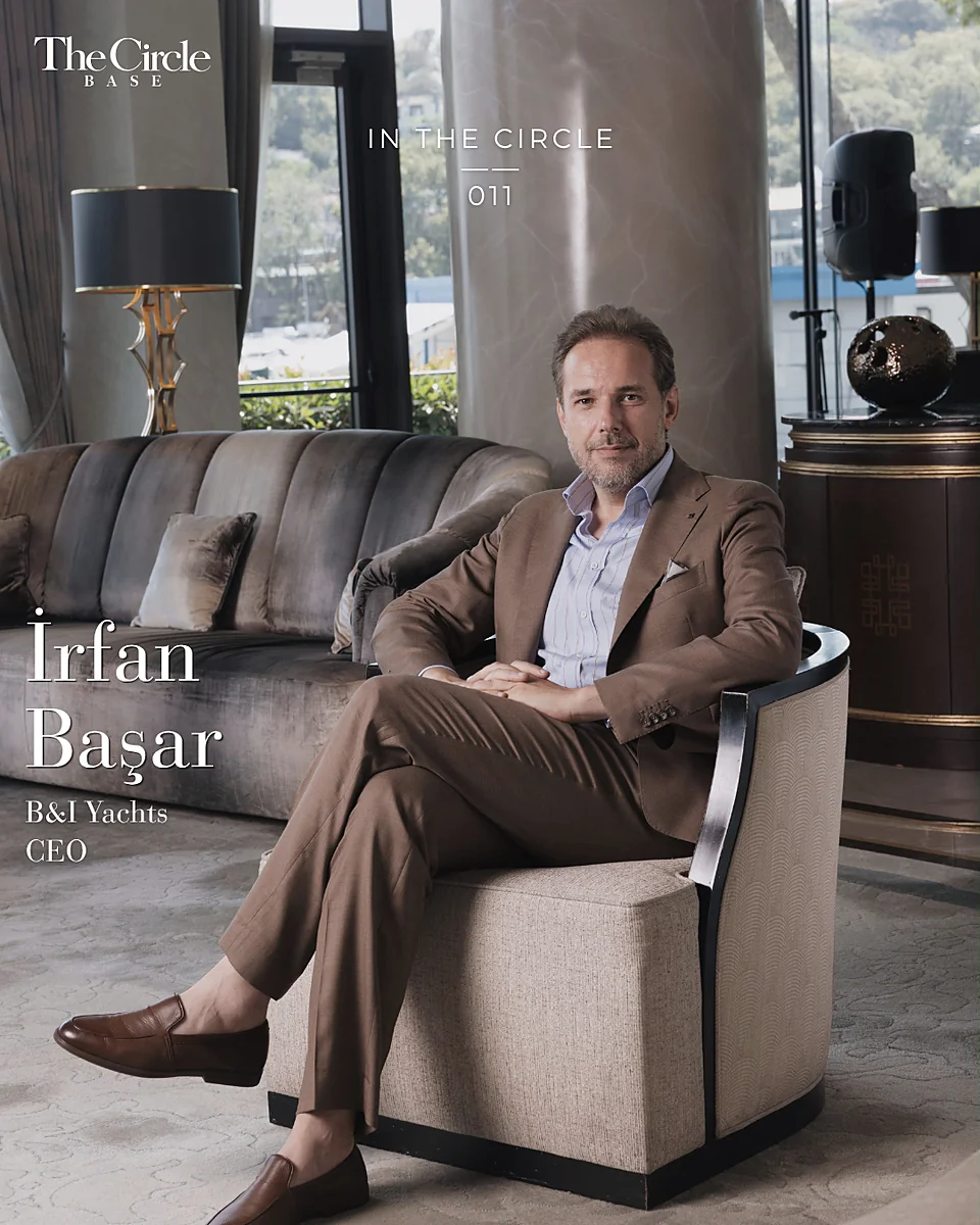 İrfan Başar seated in an armchair in a tailored suit, a yacht marina behind him